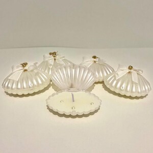 Peut inclure: Plusieurs bougies en forme de coquillage blanc. Chaque bougie a une texture nacrée et est ornée d'un ruban blanc et d'accents dorés. Une bougie est ouverte, révélant une mèche et de petites perles décoratives.