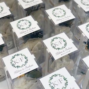 May include: Clear plastic boxes with white labels featuring a green leafy wreath with the initials "KF" and the text "Kubra & Furkan 29.01.2024".