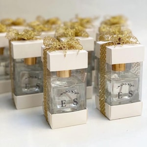May include: A set of small, clear glass bottles with gold lids and white labels. The bottles are filled with a clear liquid and have a small sprig of greenery on the label. The bottles are tied with gold ribbon and are arranged in a row on a white surface. The labels have the initials "ES" on them.