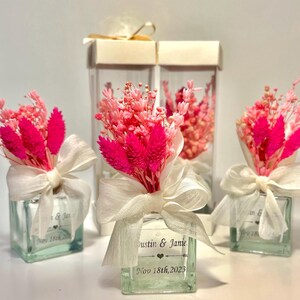 Op de afbeelding: Bruiloftfavoren in heldere glazen potten met roze gedroogde bloemen en wit lint. De potten hebben gepersonaliseerde etiketten met de namen "Dustin & Janie" en de trouwdatum "Nov 18th, 2023".
