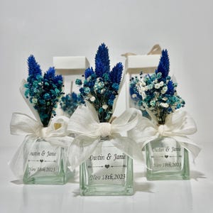 Op de afbeelding: Drie kleine glazen flesjes met gedroogde blauwe en witte bloemen, vastgebonden met een wit lint en een gepersonaliseerd etiket met de tekst "Dustin & Janie Nov 18th, 2023".