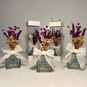 May include: Three clear glass bottles containing dried flowers and a white ribbon bow. Each bottle has a personalised label with the names "Dustin & Janie" and the date "Nov 18th, 2023".