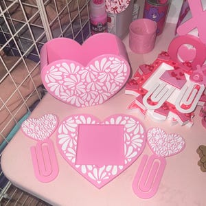 May include: A collection of pink heart-shaped desk accessories, including a pen holder, picture frame, paper clips, and decorative hearts. The items feature a white floral design. The theme is Valentine's Day or a similar romantic occasion.