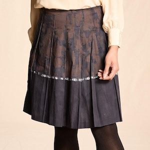 May include: A cream-colored blouse with a keyhole neckline and long sleeves paired with a pleated skirt. The skirt features a dark blue base with a brown floral pattern and a silver trim. Black tights and shoes complete the outfit.