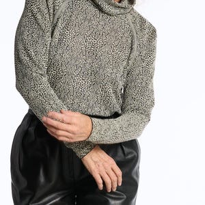 Puede incluir: Una persona con una camisa de cuello alto de manga larga con un patrón moteado en tonos grises y negros. La persona también lleva pantalones de cuero negros. El fondo es blanco liso.