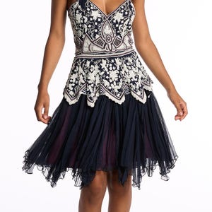 Pode incluir: Um vestido azul marinho e branco adornado com um corpete em decote em V e alças finas. O corpete apresenta intrincados padrões florais e geométricos. A saia é um design em camadas e fluido em azul marinho, com bainha com babados. Saltos pretos completam o look.