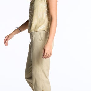 Könnte beinhalten: Ein goldfarbenes Pailletten-Top mit Flügelärmeln und eine beige Hose. Die Hose hat goldfarbene Knöpfe an der Seite des Knöchels. Das Outfit wird durch cremefarbene Slipper vervollständigt.