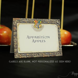 Puede incluir: Un cartel con un fondo de ladrillo y un escudo de Hogwarts que dice "Apparition Apples". El cartel está delante de una bandeja de manzanas rojas en palos.