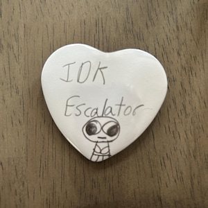 May include: White heart-shaped ceramic ornament with black marker writing that says "IDK Escalator" and a simple drawing of a person with large eyes.