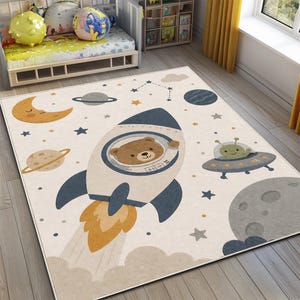 May include: A rectangular rug with a space theme. The rug features a rocket ship with a bear, an alien in a spaceship, planets, stars, and a crescent moon in shades of blue, yellow, and grey on a cream background.