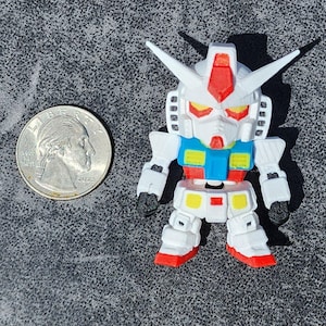 May include: A small white, blue, red, and yellow robot figurine with a pointed head crest and angular armor plating. The figure is positioned next to a standard United States quarter coin for scale, showing its compact size and detailed construction.