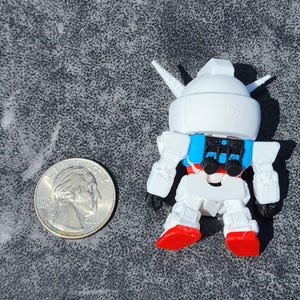 May include: A small white robot figurine with a blue backpack and red feet, positioned next to a United States quarter coin for scale. The robot features a rounded head with two pointed antennae and articulated limbs, rendered in a stylized design.