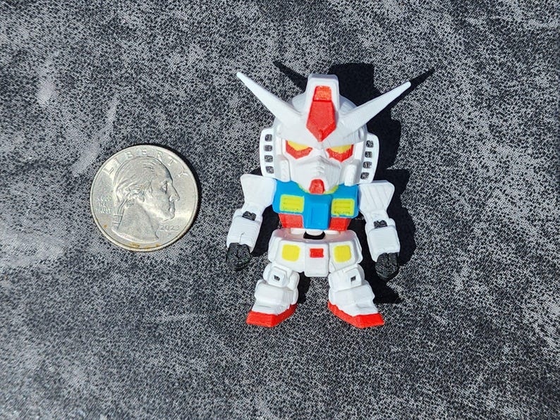 May include: A small white, blue, red, and yellow robot figurine with a pointed head crest and angular armor plating. The figure is positioned next to a standard United States quarter coin for scale, showing its compact size and detailed construction.