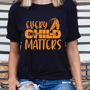 May include: Black t-shirt with the orange text "EVERY CHILD MATTERS" and a feather and arrow design. The shirt is a crew neck and has short sleeves. The shirt is worn by a person sitting in a wicker chair.