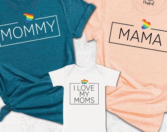 May include: Three t-shirts with a rainbow heart design and text. The first t-shirt is teal blue with the text "MOMMY". The second t-shirt is peach with the text "MAMA". The third t-shirt is white with the text "I LOVE MY MOMS".