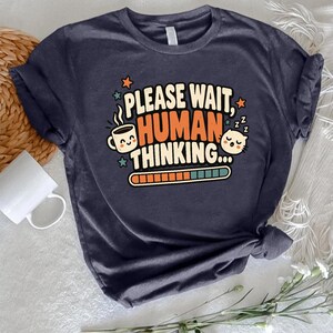 May include: A dark gray short-sleeved t-shirt featuring a graphic print that reads "PLEASE WAIT, HUMAN THINKING..." in white and orange text. The design includes a smiling coffee mug, a sleeping cat, small stars, and a loading progress bar.