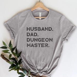 May include: A gray t-shirt with the words "HUSBAND. DAD. DUNGEON MASTER." printed in black. The shirt is hanging on a wooden hanger against a white background. A sprig of greenery is placed on the lower left side of the shirt.