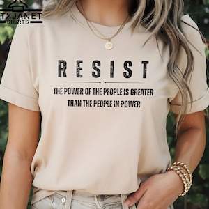 May include: Beige t-shirt with the word "RESIST" in bold, black letters. Below, the text reads "THE POWER OF THE PEOPLE IS GREATER THAN THE PEOPLE IN POWER." The shirt has short sleeves and a crew neck.