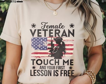May include: Beige t-shirt with the text "Female Veteran" above an American flag design and a silhouette of a soldier. Below the flag, the text reads "Touch Me And Your First Lesson Is Free."