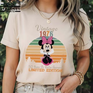 May include: Beige t-shirt featuring a Minnie Mouse graphic with a pink bow, set against a retro sunset design. The text reads "Vintage 1976" and "50 Years Of Being Awesome Limited Edition."