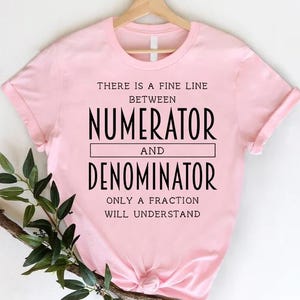 May include: A pink t-shirt with a math-themed graphic that says "There is a fine line between numerator and denominator. Only a fraction will understand."