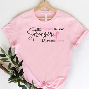 May include: A light pink t-shirt with the text "The Comeback Is Always Stronger Than The Setback" in black script and a pink ribbon graphic. The shirt is hanging on a wooden hanger.