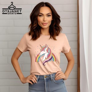K&ouml;nnte beinhalten: Pfirsichfarbenes T-Shirt mit einem bunten Einhorn-Design, Regenbogen und Sternen. Das Einhorn hat ein goldenes Horn und eine flie&szlig;ende M&auml;hne. Der Text "DTXJANET SHIRTS" befindet sich oben links.