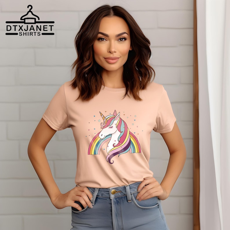 K&ouml;nnte beinhalten: Pfirsichfarbenes T-Shirt mit einem bunten Einhorn-Design, Regenbogen und Sternen. Das Einhorn hat ein goldenes Horn und eine flie&szlig;ende M&auml;hne. Der Text "DTXJANET SHIRTS" befindet sich oben links.
