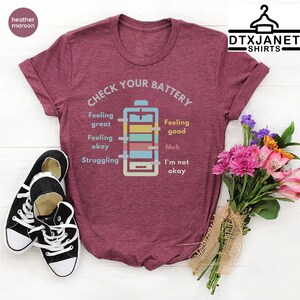 May include: Heather maroon t-shirt with the text "Check Your Battery" and a battery graphic indicating different emotional states. Black canvas sneakers and a bouquet of flowers are also in the image.