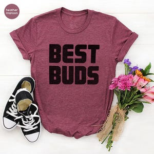 May include: Heather maroon t-shirt with the text "BEST BUDS" in black block letters. A pair of black and white canvas sneakers and a bouquet of pink and purple flowers are next to the shirt.
