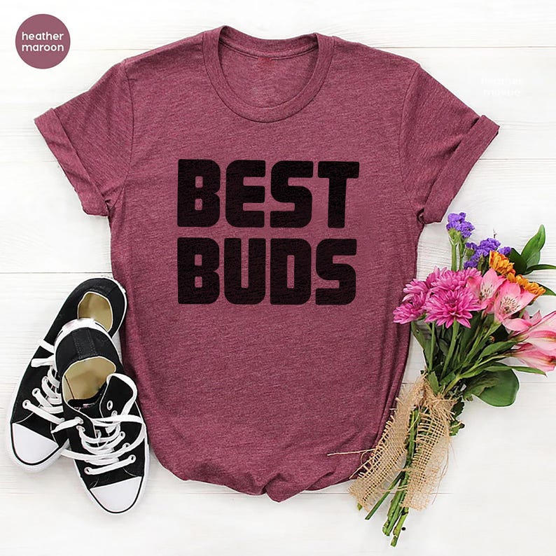 May include: Heather maroon t-shirt with the text "BEST BUDS" in black block letters. A pair of black and white canvas sneakers and a bouquet of pink and purple flowers are next to the shirt.