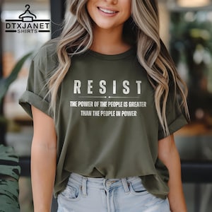 May include: Olive green t-shirt with the word "RESIST" in white block letters. Below the word "RESIST" is the text "THE POWER OF THE PEOPLE IS GREATER THAN THE PEOPLE IN POWER". The shirt has rolled-up sleeves.