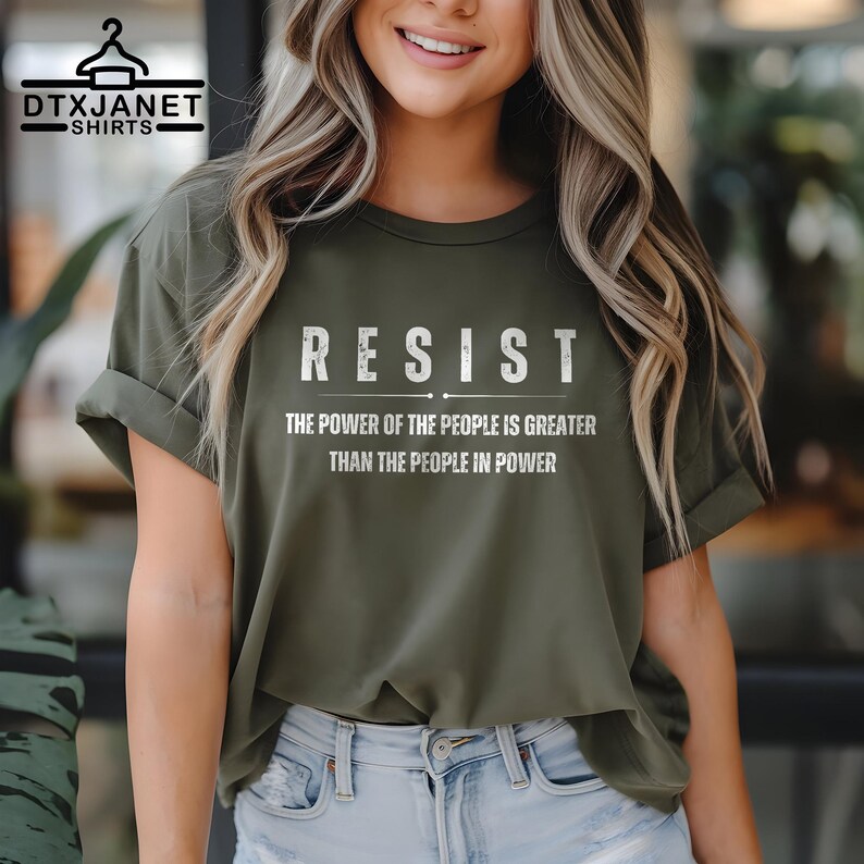 May include: Olive green t-shirt with the word "RESIST" in white block letters. Below the word "RESIST" is the text "THE POWER OF THE PEOPLE IS GREATER THAN THE PEOPLE IN POWER". The shirt has rolled-up sleeves.
