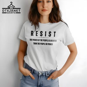 May include: White t-shirt with the word "RESIST" in bold black letters. Below the word "RESIST" is the text "THE POWER OF THE PEOPLE IS GREATER THAN THE PEOPLE IN POWER". The shirt also has the logo "DTXJANET SHIRTS" in the upper left corner.