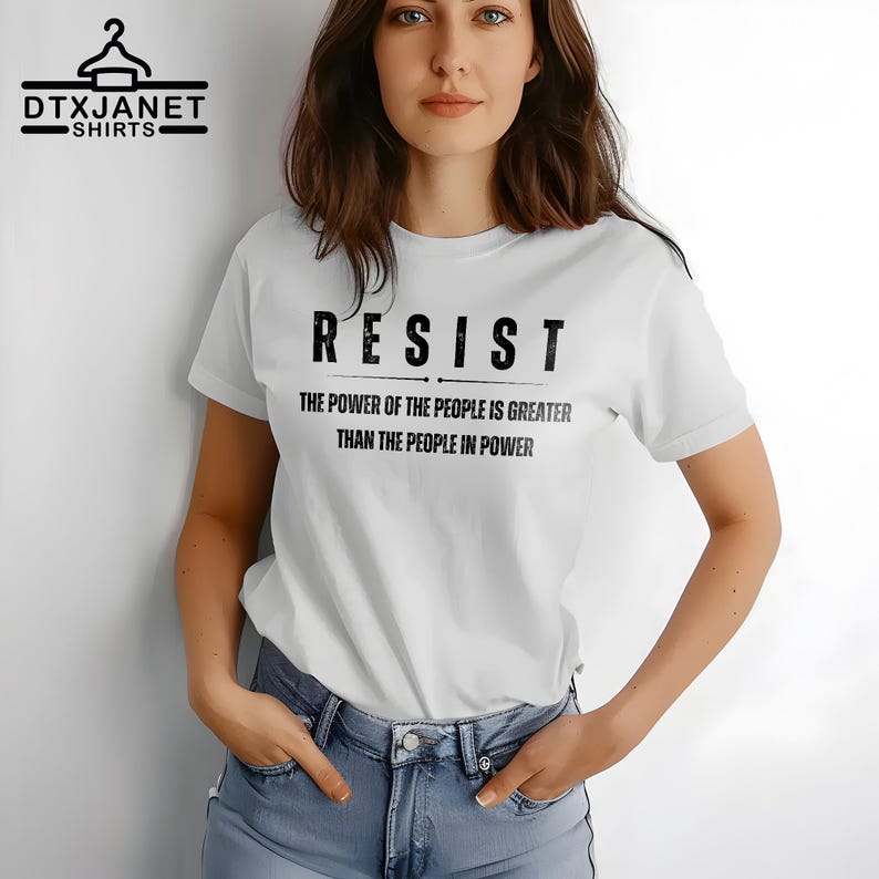 May include: White t-shirt with the word "RESIST" in bold black letters. Below the word "RESIST" is the text "THE POWER OF THE PEOPLE IS GREATER THAN THE PEOPLE IN POWER". The shirt also has the logo "DTXJANET SHIRTS" in the upper left corner.
