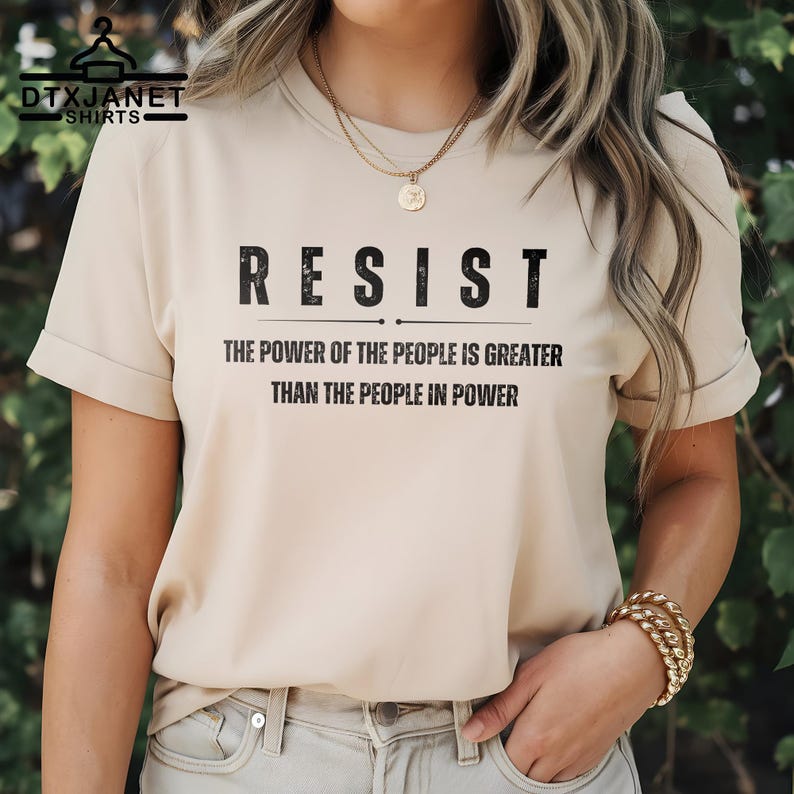May include: Beige t-shirt with the word "RESIST" in bold, black letters. Below, the text reads "THE POWER OF THE PEOPLE IS GREATER THAN THE PEOPLE IN POWER." The shirt has short sleeves and a crew neck.