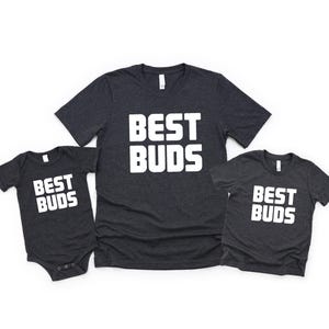 May include: Three dark gray t-shirts and a onesie with the text "BEST BUDS" in bold white letters. The shirts are short-sleeved and have a crew neck. The onesie has short sleeves and snaps at the bottom.
