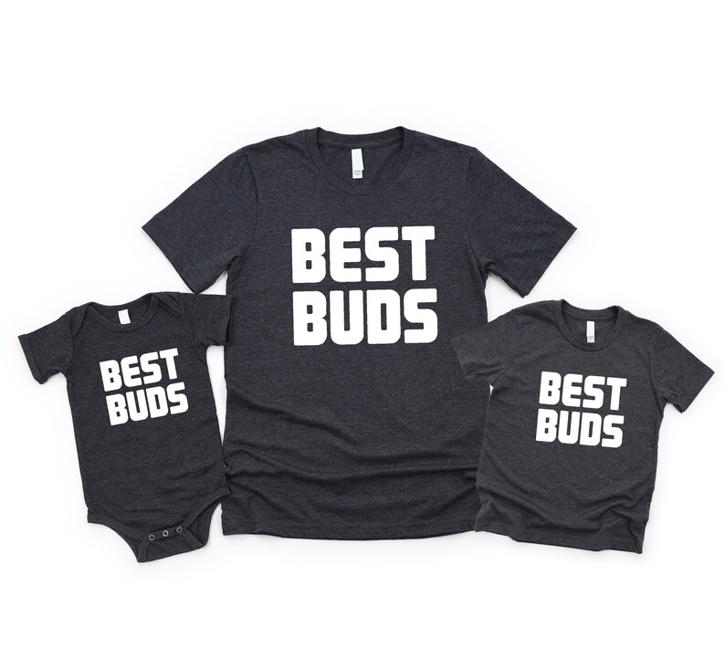 May include: Three dark gray t-shirts and a onesie with the text "BEST BUDS" in bold white letters. The shirts are short-sleeved and have a crew neck. The onesie has short sleeves and snaps at the bottom.