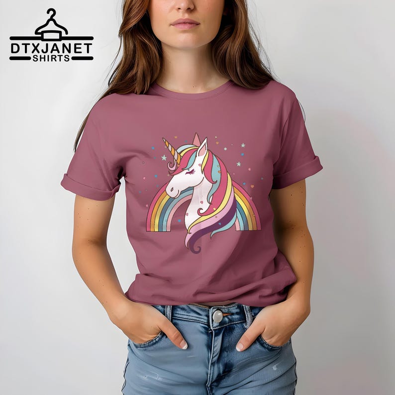 K&ouml;nnte beinhalten: Mauvefarbenes T-Shirt mit einem bunten Einhorn-Design, Regenbogen und Sternen. Das Einhorn hat ein goldenes Horn und eine flie&szlig;ende M&auml;hne. Das T-Shirt hat hochgekrempelte &Auml;rmel. Der Text "DTXJANET SHIRTS" steht oben.