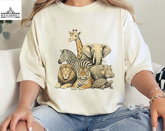 May include: A cream-colored t-shirt featuring a detailed graphic print of various wild animals including a giraffe, elephant, zebra, lion, tiger, hippopotamus, and a tortoise arranged in a cluster on the front center of the garment.