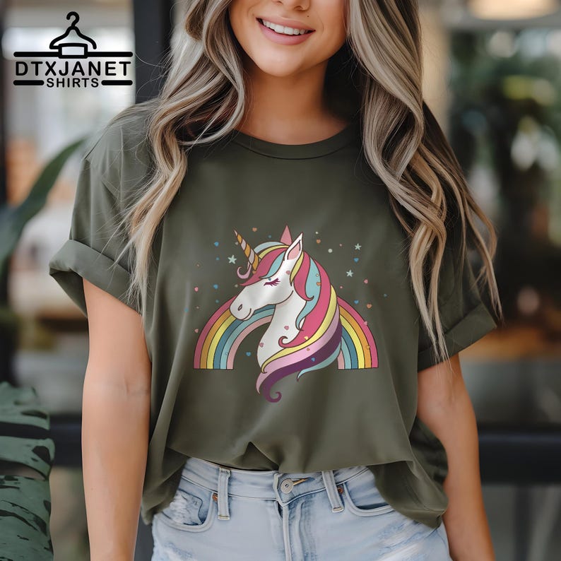 K&ouml;nnte beinhalten: Olivgr&uuml;nes T-Shirt mit einem bunten Einhorn-Design. Das Einhorn hat ein wei&szlig;es Gesicht, ein goldenes Horn und eine farbenfrohe M&auml;hne. Ein Regenbogen und Sterne erg&auml;nzen das Design. Der Text "DTXJANET SHIRTS" befindet sich oben links.