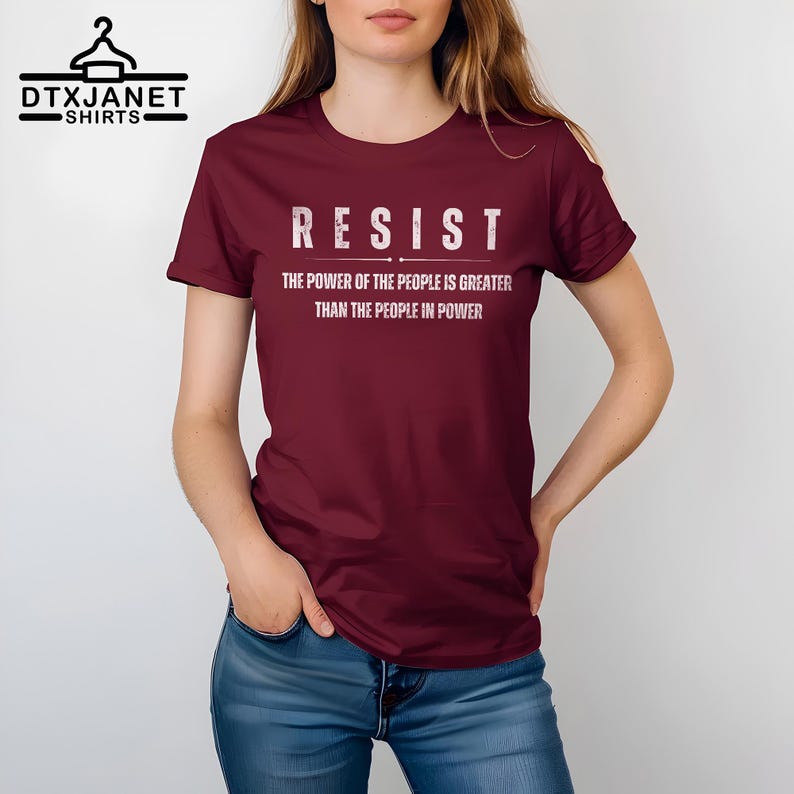 May include: A maroon t-shirt with the word "RESIST" in white block letters. Below the word, the text reads "THE POWER OF THE PEOPLE IS GREATER THAN THE PEOPLE IN POWER". The shirt is being worn by a person.