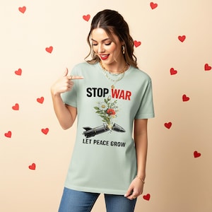 May include: A light green t-shirt with the words "STOP WAR" in black and red, and "LET PEACE GROW" in black. The graphic shows a missile with flowers growing out of it. The person is wearing blue jeans and pointing at the shirt.