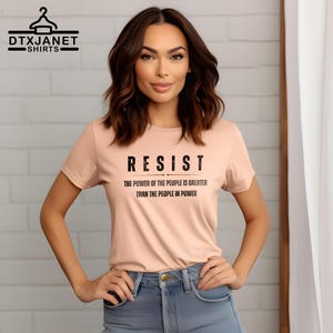 May include: A light peach-colored t-shirt with the word "RESIST" in bold black letters. Below, the text reads "THE POWER OF THE PEOPLE IS GREATER THAN THE PEOPLE IN POWER." The shirt is a crew neck style.