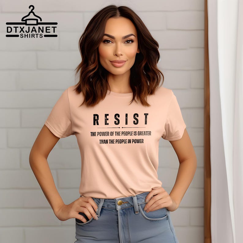 May include: A light peach-colored t-shirt with the word "RESIST" in bold black letters. Below, the text reads "THE POWER OF THE PEOPLE IS GREATER THAN THE PEOPLE IN POWER." The shirt is a crew neck style.