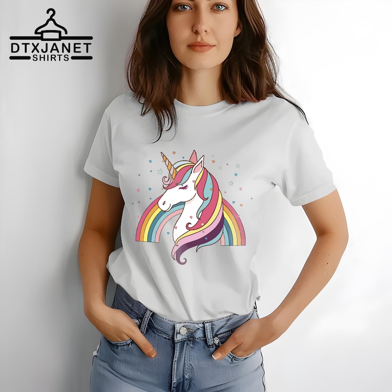 K&ouml;nnte beinhalten: Wei&szlig;es T-Shirt mit einem bunten Einhorn-Design, Regenbogen und Sternen. Das Einhorn hat ein goldenes Horn, eine rosa und blaue M&auml;hne und einen lila Schwanz. Der Text "DTXJANET SHIRTS" ist schwarz gedruckt.
