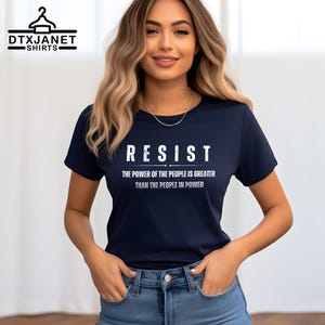 May include: Navy blue t-shirt with the word "RESIST" in white block letters. Below the word "RESIST" is the text "THE POWER OF THE PEOPLE IS GREATER THAN THE PEOPLE IN POWER". The shirt is a crew neck style.