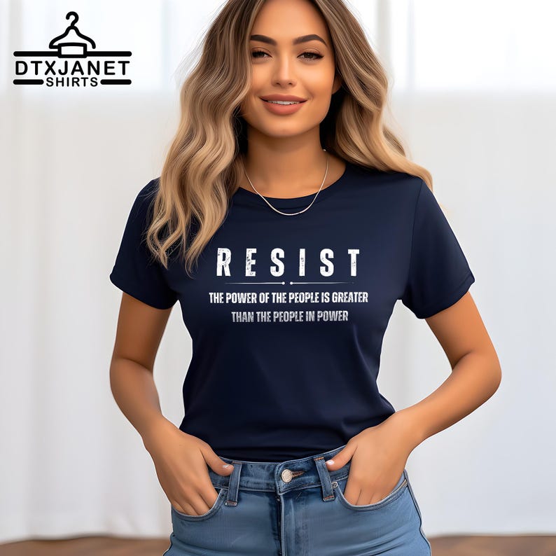 May include: Navy blue t-shirt with the word "RESIST" in white block letters. Below the word "RESIST" is the text "THE POWER OF THE PEOPLE IS GREATER THAN THE PEOPLE IN POWER". The shirt is a crew neck style.