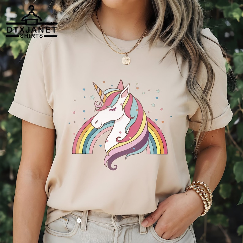 K&ouml;nnte beinhalten: Beiges T-Shirt mit einem farbenfrohen Einhorn-Design, einschlie&szlig;lich Regenbogen, Sternen und Herzen. Das Einhorn hat ein goldenes Horn und eine flie&szlig;ende M&auml;hne in Pink, Gelb und Blau. Der Text "DTXJANET SHIRTS" steht oben links.