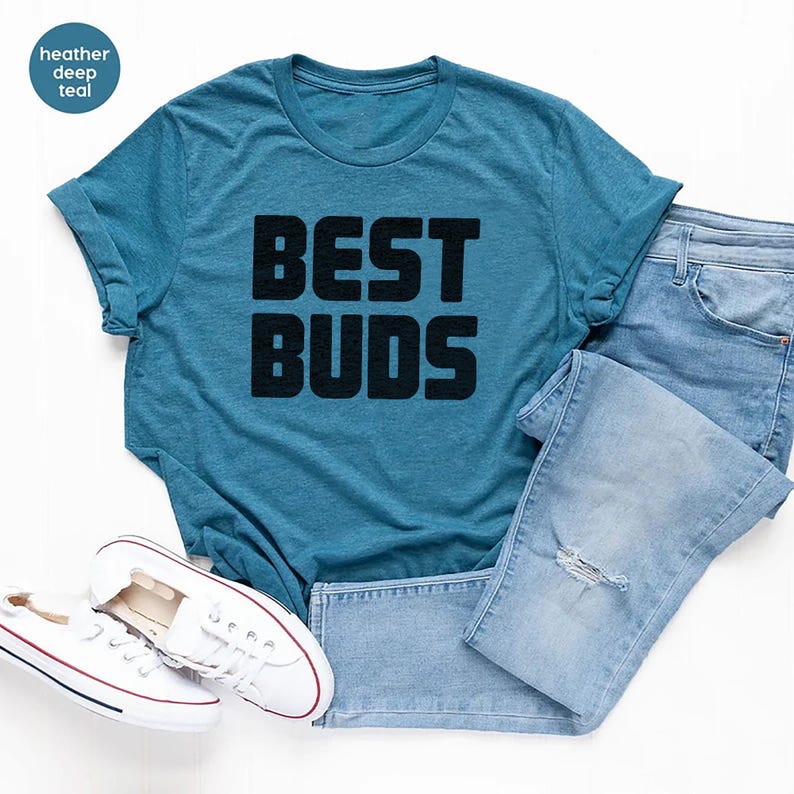 May include: A teal t-shirt with the words "BEST BUDS" in bold black letters. The shirt is paired with light blue denim jeans and white sneakers with red accents. The shirt is made of a soft material.